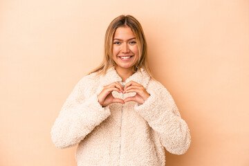Young caucasian woman isolated on beige background smiling and showing a heart shape with hands.