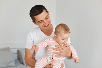 Smiling handsome young adult father with his toddler baby girl wearing striped sleeper, enjoying taking care of his cute kid, playing together with daughter at home.