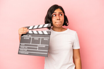 Young latin woman holding clapperboard isolated on pink background confused, feels doubtful and...