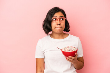 Young latin woman holding cereals isolated on pink background confused, feels doubtful and unsure.