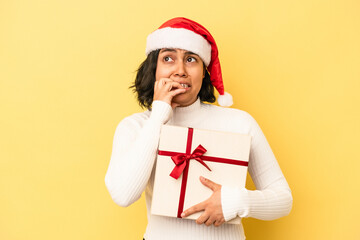 Young latin woman celebrating christmas holding a gift isolated on yellow background biting fingernails, nervous and very anxious.