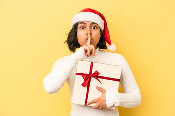 Young latin woman celebrating christmas holding a gift isolated on yellow background keeping a secret or asking for silence.