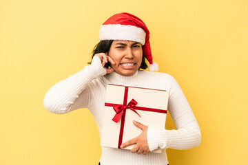 Young latin woman celebrating christmas holding a gift isolated on yellow background covering ears with hands.