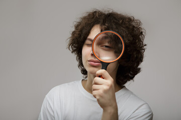 Happy young teenage man with curly hair wearing glasses holds a magnifying glass in his hands. Gray background