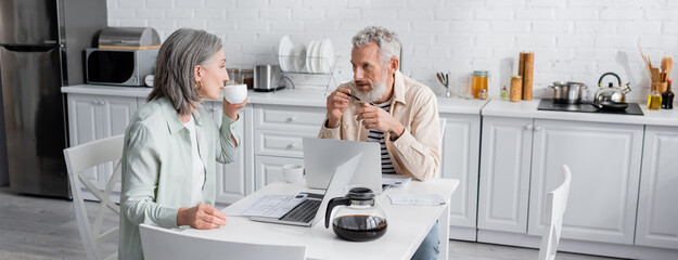 Mature man talking to wife with coffee near laptops and bills in kitchen, banner.