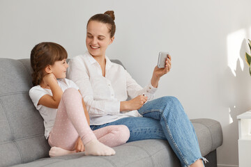 Positive people posing indoor at home, having video call, woman holding smart phone in hand and looking at her daughter, mother blogger broadcasting livestream, talking with subscribers.