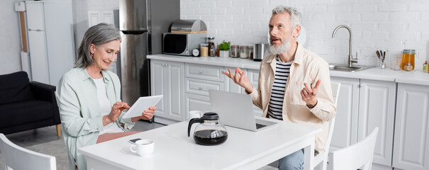 Smiling woman using digital tablet near pensive mature husband and coffee in kitchen, banner.