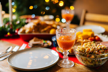 Close-up of glass with fresh orange juice and delicious vegetable salad on feast Christmas dinner table during holiday friendly family party, on blurred background of xmas celebration bokeh lights.