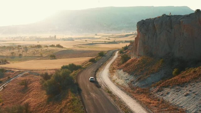 Drone camera follows white car moving along epic beautiful mountain and field sunset landscape road in summer Cappadocia