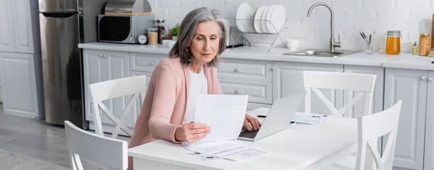 Grey haired mature woman holding bills near laptop in kitchen, banner.