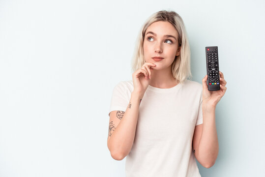 Young caucasian woman holding a tv controller isolated on blue background looking sideways with doubtful and skeptical expression.