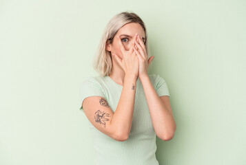 Young caucasian woman isolated on green background blink through fingers frightened and nervous.