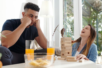 Pretty young woman laughing celebrating victory in board game when frustrated husband looking at tower of wooden blocks