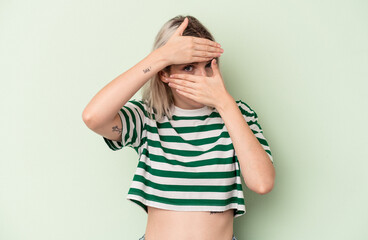 Young caucasian woman isolated on green background blink at the camera through fingers, embarrassed covering face.