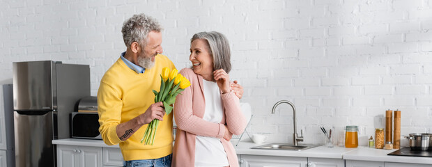 Smiling mature man hugging wife and holding yellow tulips in kitchen, banner.