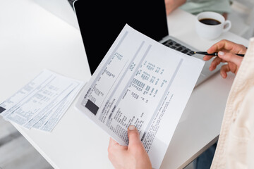 Fototapeta premium Cropped view of man holding bills near laptop and blurred coffee at home.