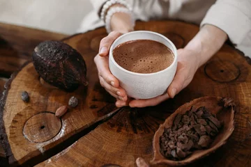 Fotobehang Hot handmade ceremonial cacao in white cup. Woman hands holding craft cocoa, top view on wooden table. Organic healthy chocolate drink prepared from beans, no sugar. Giving cup on ceremony, cozy cafe © ninelutsk