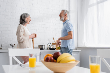 Side view of cheerful mature couple talking near stove in kitchen.