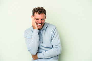 Young caucasian man isolated on green background laughs happily and has fun keeping hands on stomach.