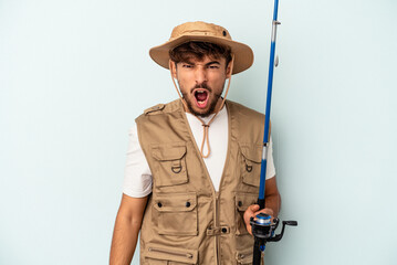 Young mixed race fisherman holding a rod isolated on blue background screaming very angry and aggressive.