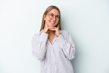 Young caucasian woman isolated on blue background keeps hands under chin, is looking happily aside.