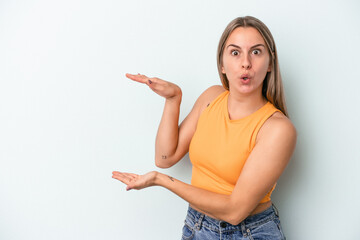 Young caucasian woman isolated on blue background shocked and amazed holding a copy space between hands.