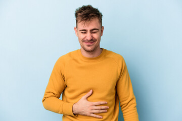 Young caucasian man isolated on blue background touches tummy, smiles gently, eating and satisfaction concept.