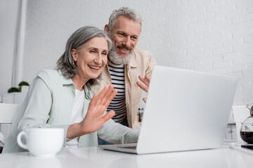 Mature couple having video chat on laptop near blurred cup of coffee at home.