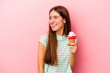 Young caucasian woman holding an ice cream isolated on pink background looks aside smiling, cheerful and pleasant.