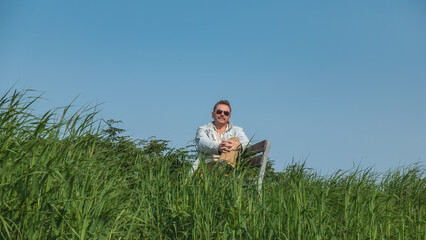Naklejka na ściany i meble A man in dark glasses is sitting resting on a bench among the lush tall green grass. Legs crossed. There's a smile on his face. In the background is a clear blue sky. A sunny summer day. Kamchatka