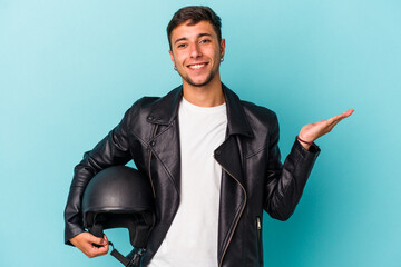 Young biker man holding helmet isolated on blue background  showing a copy space on a palm and...