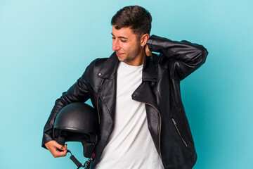 Young biker man holding helmet isolated on blue background  touching back of head, thinking and...