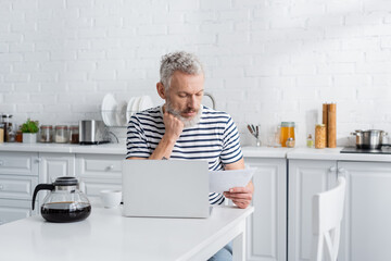 Middle aged man looking at document near coffee and laptop in kitchen.