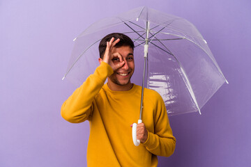 Young caucasian man holding umbrella isolated on purple background  excited keeping ok gesture on...