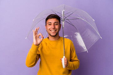 Young caucasian man holding umbrella isolated on purple background  cheerful and confident showing ok gesture.