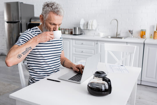 Middle aged man drinking coffee and using laptop near papers in kitchen.