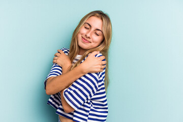 Young caucasian blonde woman isolated on blue background  hugs, smiling carefree and happy.