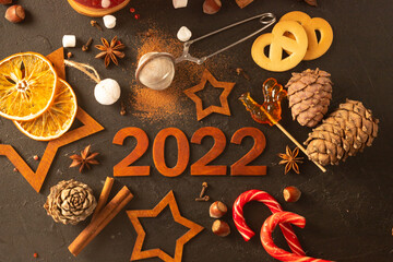 Wooden brown numbers 2022 on a dark background. There are stars carved out of wood, spices, scattered cocoa and a mug with marshmallows around