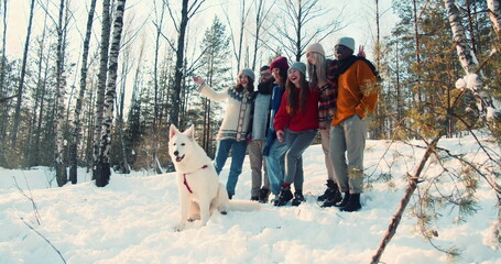 Winter vacation concept. Happy group of multiethnic friends and dog posing in sunny snowy forest together slow motion.