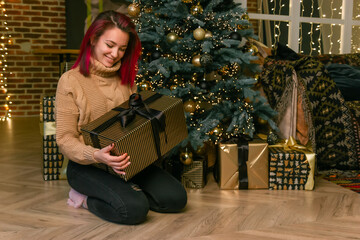Obraz na płótnie Canvas Dreamy charming young lady in a sweater with a gift in her hands on the floor against the background of a Christmas tree with gifts