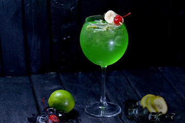 green alcoholic cocktail with ice decorated with cherry and banana on a skewer on a black background macro photo
