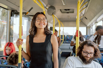 Smiling, attractive student in strange city, moving by public transport, girl is holding on to railing, looking out window with joy, around her other passengers traveling by bus to work, school