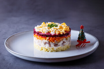 Herring salad under a fur coat. Traditional Russian multilayered salad from herring, beets, potatoes, carrots and eggs. Close-up, grey background, miniature Christmas tree.