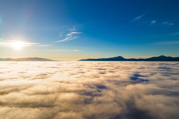 Aerial view from above of white puffy clouds at yellow sunset and distant mountains