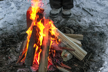 Female human foots in hiking boots by winter campfire on snow at dusk campsite in wild, bright red...