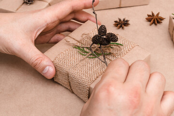 Obraz premium Cristmas decoration of gifts in eco-style,male hands close-up.Gift boxes are wrapped in craft paper,decorated with thuja leaves and cones.Christmas,New Year and eco-friendly concept.