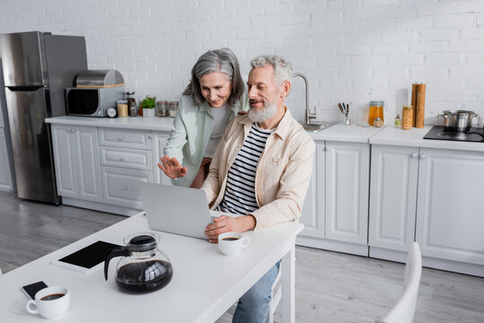 Smiling mature couple having video call on laptop near devices and coffee in kitchen.
