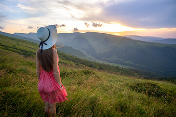 Young happy woman traveler in red dress standing on grassy hillside on a windy evening in summer mountains enjoying view of nature at sunset