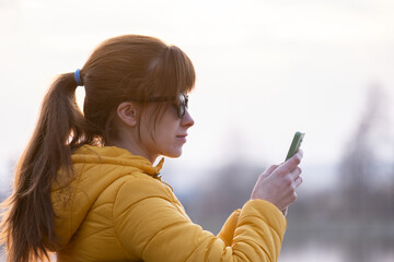 Young pretty woman sitting on a park bench browsing her smartphone outdoors on warm autumn day