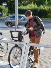 Obraz premium Young man is renting bicycle by smartphone application. Eco-friendly urban transport for rent. Healthy lifestyle in modern town. Autumn sunset.